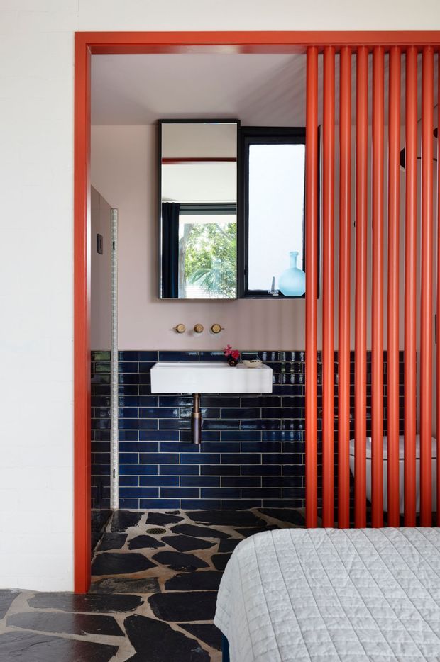 A compact bathroom viewed directly from a bedroom, with a wall-mounted white basin, dark navy-blue rectangular tiled walls, and a black irregular stone-look floor. A bold red-orange doorway frame and vertical slatted screen partially separate the bathroom from the bedroom. A black-framed mirror and window sit above the basin, with warm pale walls and a visible bed in the foreground.