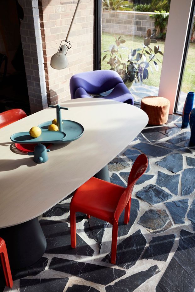 A sunlit dining area with a large pale oval dining table, sculptural dark pedestal base, colorful red dining chairs, and a purple lounge chair beside full-height glazing. The room has an exposed pale brick wall, a distinctive irregular dark stone floor with light grout, and a contemporary pendant lamp. A small tray with fruit and a blue bottle add accents, while a landscaped courtyard is visible through the windows.