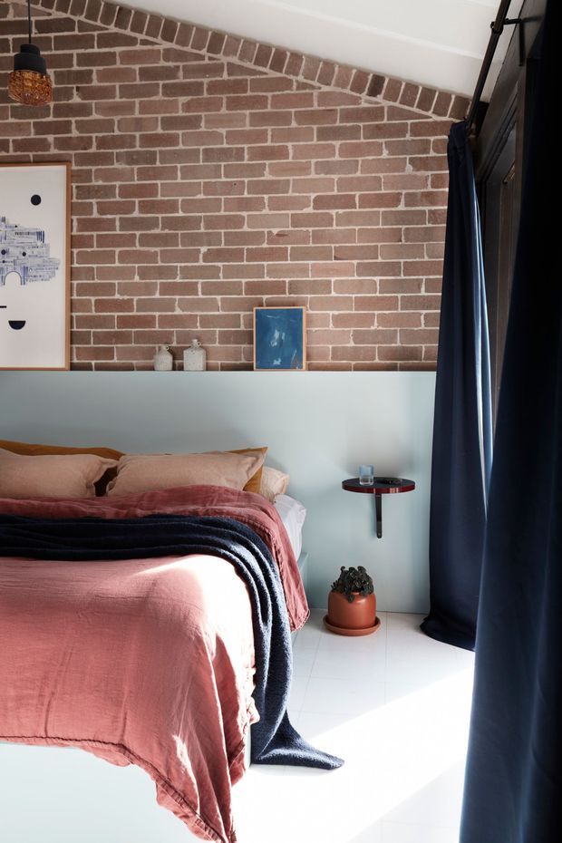 A stylish bedroom with a partially exposed red-brown brick feature wall above a pale blue painted lower wall. The room includes a bed dressed in dusty pink linen and a dark navy throw, deep navy curtains, white tiled flooring, a small round wall-mounted bedside table, ceramic accessories, framed artwork, and a potted plant. Strong sunlight creates a bright, high-contrast atmosphere.