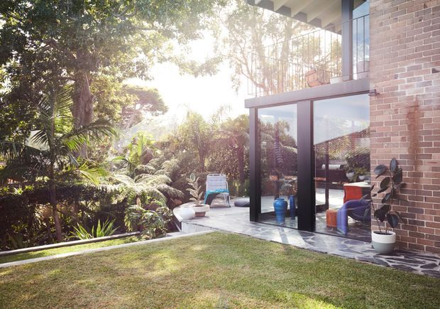A sunlit modern brick house opening onto a landscaped garden terrace. The image shows large floor-to-ceiling glazed doors and windows, a dark-framed upper balcony, a covered roof edge, and an outdoor seating area visible through the glazing. Dense tropical planting surrounds the lawn and paved terrace.