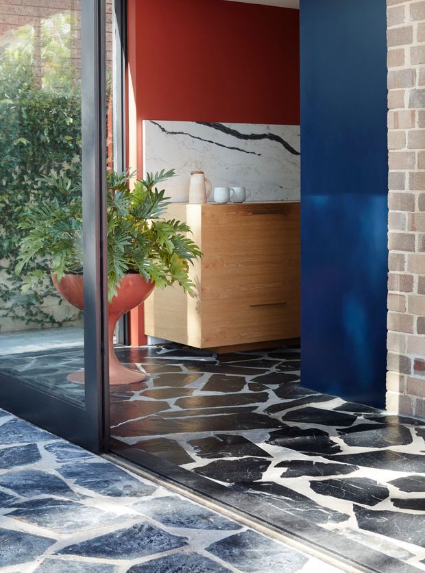 A richly coloured contemporary entryway viewed through an open glazed door. The space features an irregular dark-and-white stone floor extending inside and outside, a deep red wall, a bold navy-blue door or wall panel, exposed pale brick, and a warm timber console with a dramatic white veined stone backsplash. A large potted plant and ceramic vessels add natural and decorative accents.