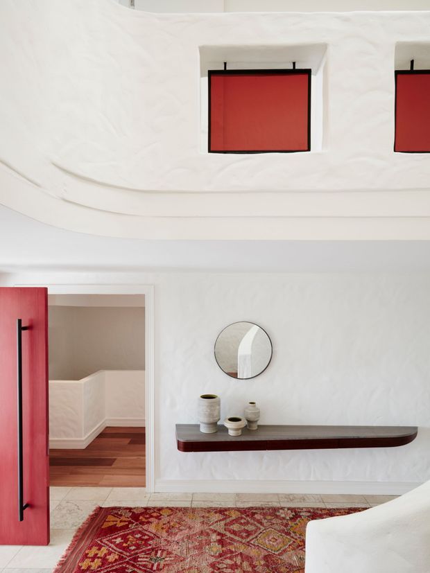 A bright, sculptural interior entry or living space with white organically textured walls and a curved white ceiling featuring recessed red geometric panels. A bold red door opens toward a small adjoining area with dark timber flooring. The main wall has a floating dark timber console, round mirror, and ceramic vessels, with a patterned red rug and part of a white sofa in the foreground. The space combines minimalist built forms with strong red accents and warm wood details.