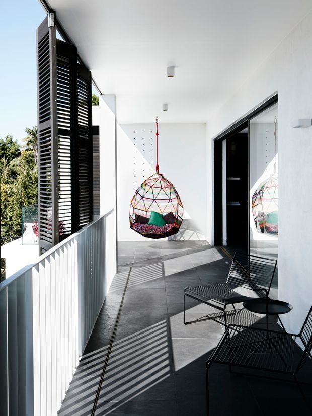 A bright, modern covered outdoor terrace or balcony with dark tiled flooring, white walls and ceiling, black-framed glazing, and a black louvred privacy screen. A colourful hanging egg chair provides a focal point, while black wire-frame lounge chairs and a small round table form a seating area. Strong sunlight casts crisp striped shadows from the screen and balustrade across the floor and walls, with greenery visible beyond the railing.