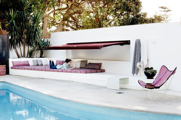 A bright outdoor poolside lounge with a long built-in white rendered bench and recessed alcove, striped upholstered cushions, a matching striped sling chair, and a narrow dark red wall-mounted canopy or shelf. Pale paving surrounds the turquoise swimming pool, with tropical planting along the rear boundary wall. The scene has a clean, relaxed, resort-like character.