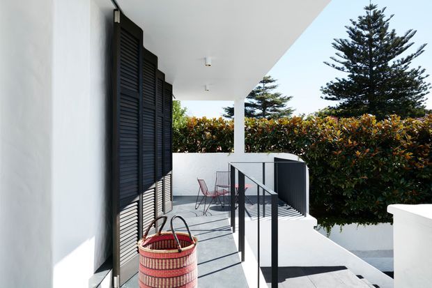 Bright exterior balcony or terrace beside a white modern building, featuring dark louvred shutters, a covered overhang, concrete paving, black metal balustrades, white rendered walls, and a small outdoor dining setting. Dense clipped hedging and mature evergreen trees frame the space.