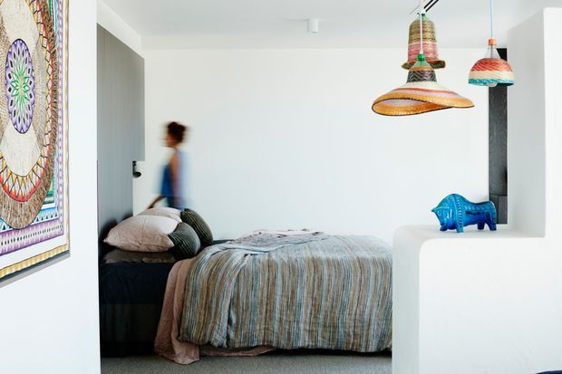 A bright, compact bedroom with a low bed dressed in layered neutral and blue-grey bedding, a dark upholstered or timber-toned headboard, and white walls. A large colourful patterned artwork hangs on the left wall, while three distinctive woven pendant lights hang above the room. A white built-in partition or ledge in the foreground displays a small blue decorative elephant. The space has a playful, eclectic, colourful character with clean contemporary architectural finishes.