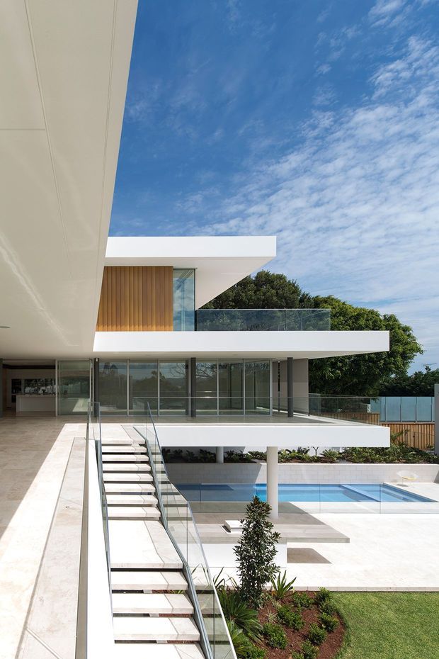 A bright multi-level contemporary residence viewed from an elevated terrace or balcony, featuring crisp white cantilevered slabs, extensive floor-to-ceiling glazing, slim dark frames, warm vertical timber wall cladding, and a glass balustrade. An open stair descends alongside a landscaped courtyard and rectangular swimming pool with pale stone paving. The composition emphasizes clean geometric forms, outdoor-indoor connection, and a vivid blue sky.