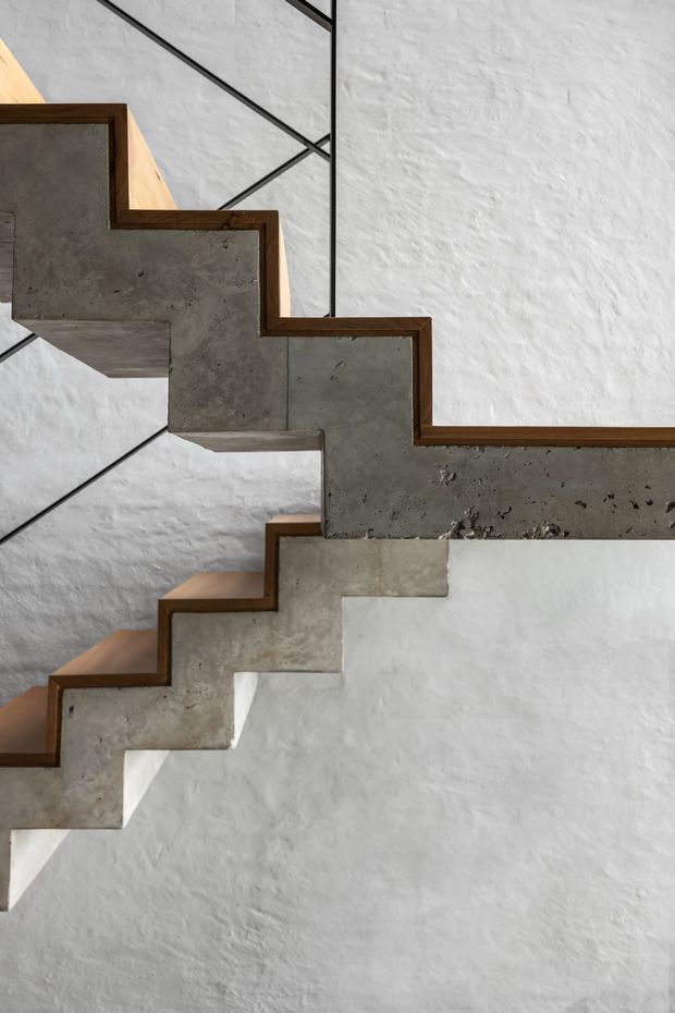 Close-up architectural view of a sculptural zigzag stairway with exposed concrete stringers and treads edged in dark timber. The staircase is set against a textured white plaster wall with slim black metal balustrade elements, creating a striking geometric composition.
