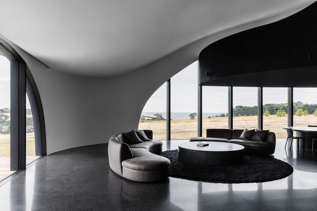 A dramatic contemporary living room with sculptural curved white walls and a sweeping ceiling, expansive floor-to-ceiling glazing, and dark polished flooring. The space is furnished with low black and charcoal sofas, a large round black coffee table, and a textured dark rug. A rural landscape is visible outside through the glazing, while a dark upper volume and dining area extend to the right.