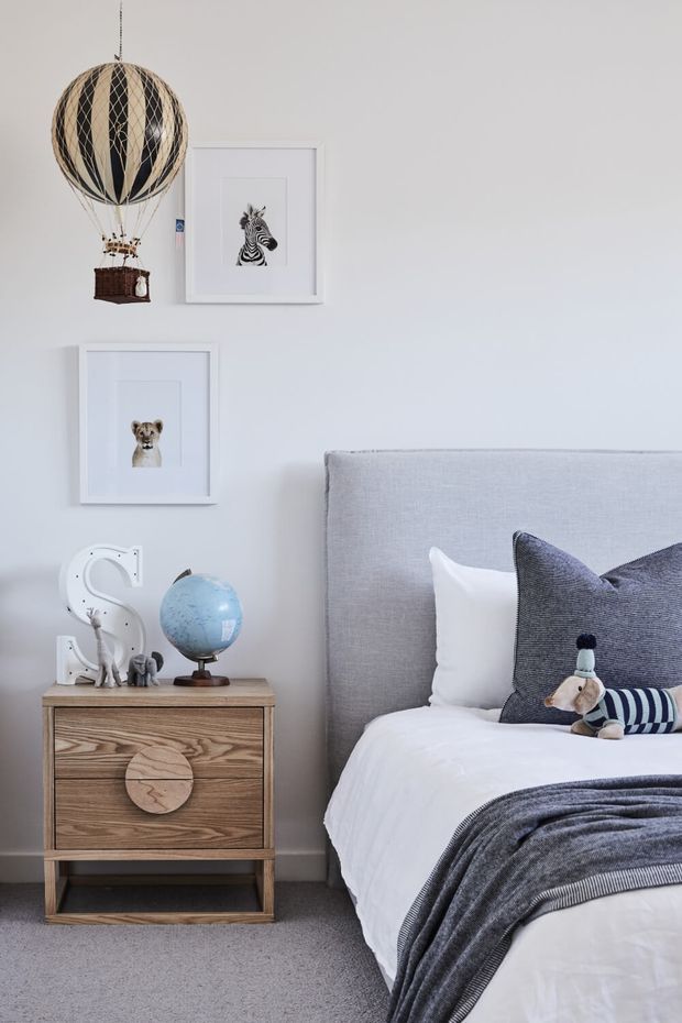 A softly styled children's bedroom featuring a light grey upholstered bed, white bedding, layered charcoal and striped cushions, and a coordinating throw. A timber bedside table holds a blue globe, decorative letter, and small animal figurines. Framed zebra and lion artwork and a whimsical hanging hot-air-balloon ornament add playful character. The room has pale walls, grey carpet, and a calm neutral palette with blue accents.