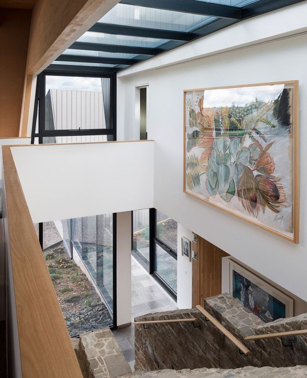 A bright, double-height stairwell and landing with a glazed roof bringing in natural light. The space features white plaster walls, dark-framed floor-to-ceiling glazing, warm timber wall and stair elements, and rugged irregular stone stair and landing surfaces. A large botanical artwork is mounted on the upper wall, while a lower-level entry or living area is visible below.