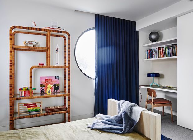 Bright, contemporary bedroom with a built-in home-office nook. The room features white walls and ceiling, a round window partly concealed by a deep navy pleated curtain, open white shelving filled with books, a compact white desk, and a timber chair. A sculptural rattan or cane shelving unit displays decorative objects and children's items. The bed occupies the foreground with a textured blue-grey throw and light bedding, creating a playful yet refined interior.