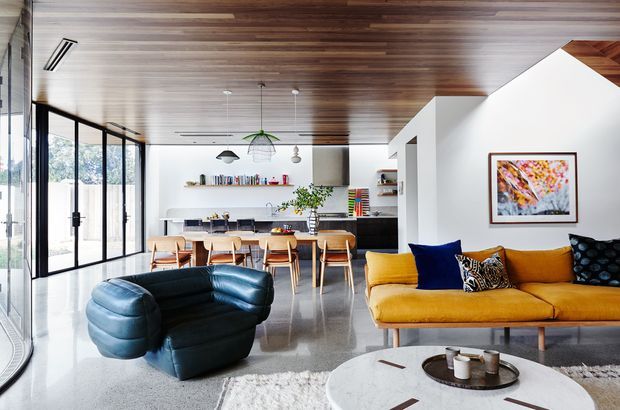 Open-plan contemporary living and dining area with a kitchen at the rear. The space features a striking timber-lined ceiling, full-height black-framed glazing along one side, polished grey concrete flooring, white walls, a mustard yellow sofa, dark blue sculptural lounge chair, timber dining table and chairs, pendant lighting, and colourful artwork. The room is bright, spacious, and visually connected to the outdoors.