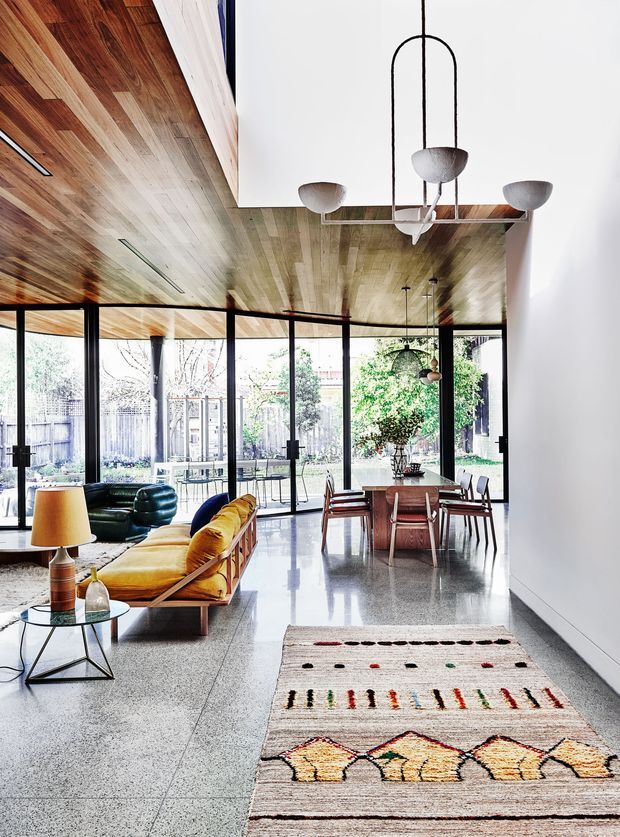 Bright open-plan modern living and dining area with floor-to-ceiling glazing, a polished terrazzo floor, warm timber-lined ceiling, and a distinctive suspended multi-light pendant. The room includes a mustard-yellow sofa, dark lounge chairs, a timber dining table with chairs, and a geometric patterned rug. The glazed rear wall opens visually to a landscaped garden.