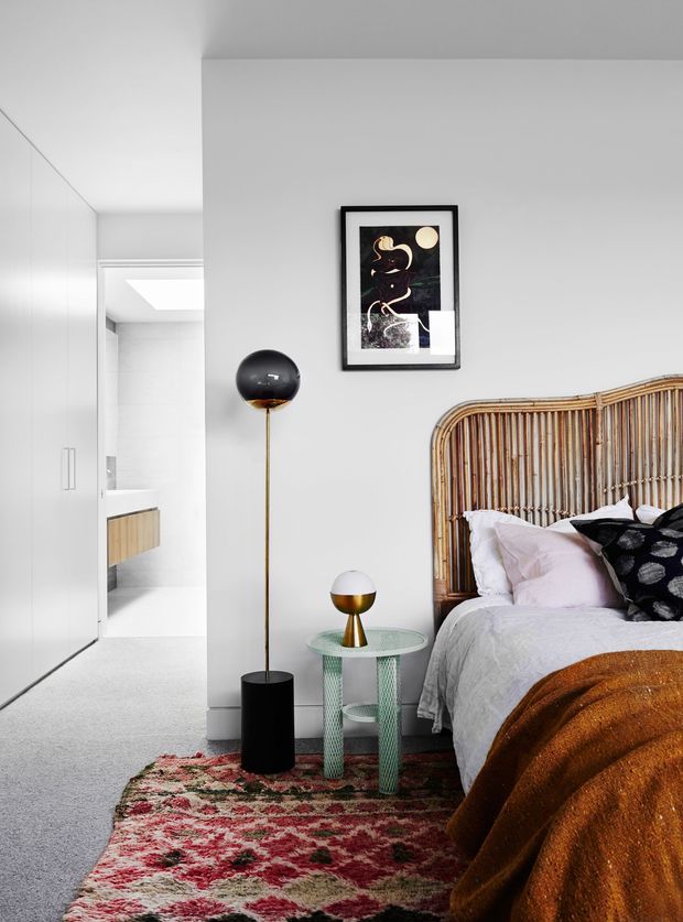 A bright, contemporary bedroom with a partially visible adjoining bathroom. The room features a tall rounded rattan or cane bedhead, white bedding, blush and black cushions, a rust-brown throw, and a richly patterned red and cream rug. A black globe floor lamp and a small sculptural table lamp sit beside a pale green perforated bedside table. White built-in wardrobes line the left side, while the background reveals a minimalist bathroom with a timber vanity and skylight.