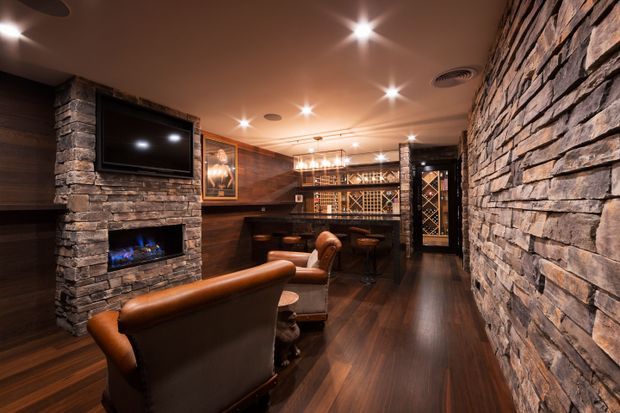 A moody, luxurious basement-style home bar and lounge with dark timber wall panelling, rich dark timber flooring, extensive rough stacked-stone cladding, a stone fireplace with gas flame, wall-mounted television, leather lounge chairs, a bar counter with stools, and illuminated wine storage at the rear. Recessed ceiling spotlights and warm pendant lighting create an intimate entertainment atmosphere.