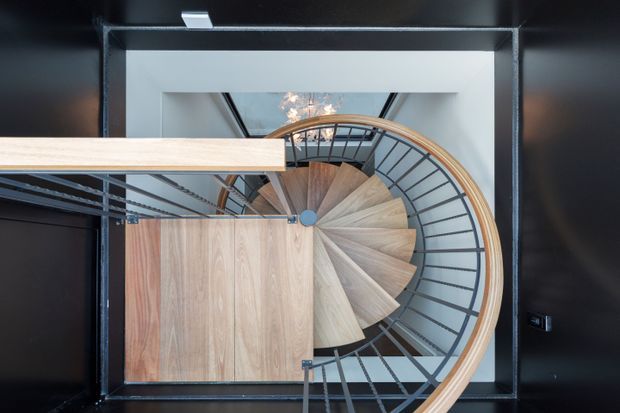 Top-down view of a compact spiral staircase within a square stairwell. The stair treads are finished in warm natural timber, while dark metal balustrades and surrounding walls create a strong black-and-wood contrast. A curved timber handrail follows the staircase perimeter, and a small skylight or ceiling opening brings light into the space.