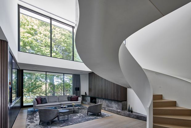 Contemporary double-height living room with extensive floor-to-ceiling glazing, a sculptural curved staircase, dark timber wall panelling, a stone fireplace wall, and a neutral seating arrangement. Lush trees are visible outside through the glazing.