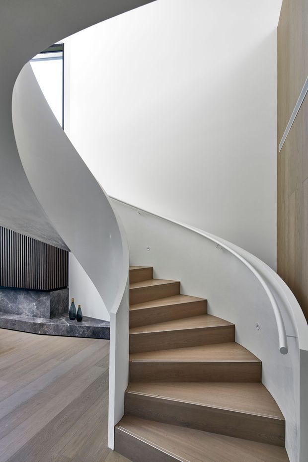 A sculptural curved staircase dominates a refined contemporary interior. Broad timber stair treads rise beside a smooth white sweeping balustrade and matching handrail, with pale timber flooring, a dark stone hearth or ledge, and vertical dark slatted wall detailing visible to the left. The space is bright, minimal, and architectural, with a tall white wall and a narrow window providing natural light.
