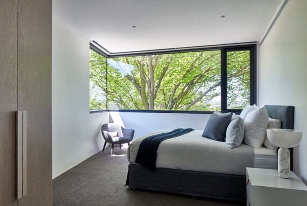 A bright, contemporary bedroom with a neatly made bed, layered white and grey bedding, a dark upholstered bed base, a sculptural bedside table and lamp, a lounge chair, and extensive black-framed corner glazing overlooking leafy trees. The room has white walls, a smooth white ceiling with recessed downlights, grey carpet, and a calm, refined palette.