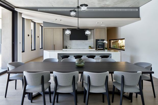 A contemporary open-plan kitchen and dining area with a long dark timber dining table surrounded by upholstered black-framed chairs. Behind it is a large white stone island with seating, integrated light timber cabinetry, a dark splashback, built-in appliances, and pendant lighting. Full-height glazing and a concrete-look ceiling contribute to the bright, refined interior.