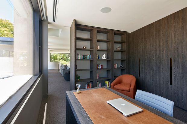 A refined home office with a large timber desk positioned beside full-height glazing. Dark timber built-in shelving and wall panelling provide substantial storage and contrast with the warm leather desk mat, rust-coloured armchair, and pale upholstered chair. The room opens toward a bright living area beyond, with integrated linear ceiling ventilation and recessed lighting.
