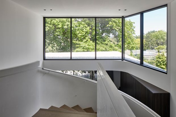 A bright contemporary stairway and landing with a descending timber staircase, white walls, a sculptural central balustrade, and extensive black-framed corner glazing. The windows overlook leafy suburban surroundings and bring abundant natural light into the double-height circulation space. A dark vertical-slatted feature wall is visible beside the lower stairwell.