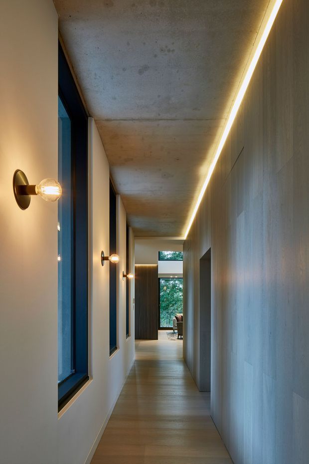 A long, narrow contemporary hallway with warm timber flooring, pale walls, a raw exposed concrete ceiling, and a continuous recessed linear light along the right-hand wall. Repeated brass wall sconces illuminate tall dark-framed windows on the left. The corridor leads toward a brighter living area with timber screening and greenery visible beyond.