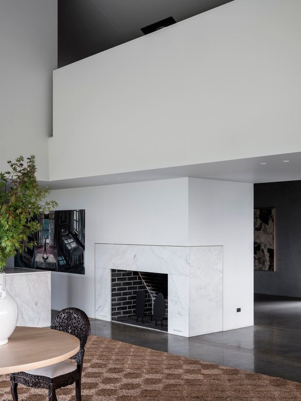A sophisticated open-plan living and dining area featuring a large sculptural white fireplace clad in pale veined stone, a black brick firebox, polished concrete flooring, and a high angular ceiling. A round timber dining table with ornate dark chairs, a patterned rug, contemporary artwork, and a large indoor plant add warmth to the otherwise monochromatic space. A dark hallway or adjoining room is visible beyond the fireplace.