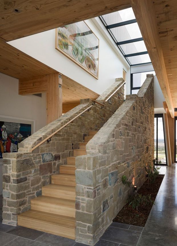 A dramatic interior stairway rises through a double-height space. The staircase is enclosed by substantial rough stone masonry walls, with pale timber stair treads and extensive exposed timber ceiling and structural framing. A large glazed roof or skylight brings daylight into the space, while tall windows reveal an exterior landscape. Artwork is mounted on the white upper wall, and a polished dark floor runs alongside a narrow planted bed.