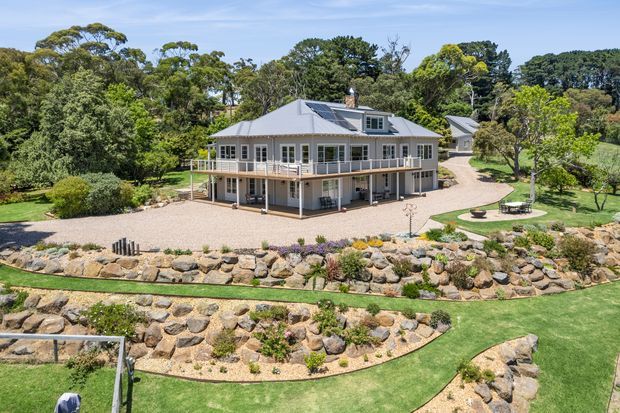 Aerial exterior view of a substantial two-storey home set within expansive landscaped grounds. The house has a light-coloured hipped metal roof, extensive white-framed glazing, wraparound upper-level verandas or balconies, and a pale rendered or weatherboard-like facade. Curving gravel paths, manicured lawns, planted rock gardens, and landscaped retaining areas surround the building.