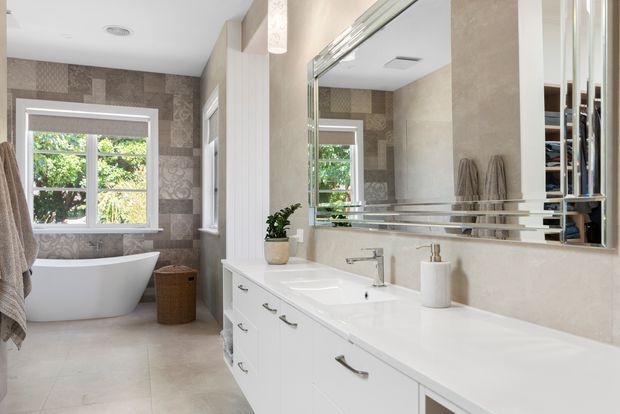 Bright, refined bathroom with a long white double vanity, extensive mirrored wall, freestanding bathtub, patterned feature wall tiles, pale stone-look surfaces, and a large window overlooking greenery. The room has a clean traditional-contemporary feel with brushed metal hardware and soft neutral finishes.