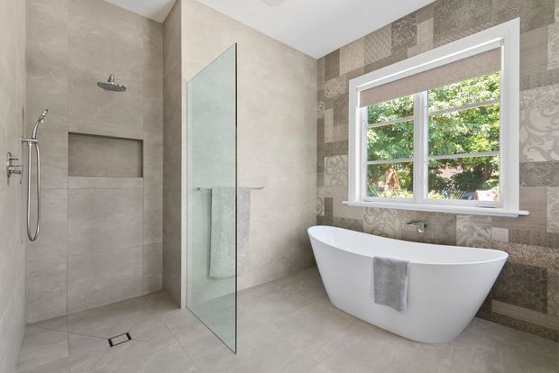 Contemporary bathroom featuring a walk-in tiled shower with recessed niche, frameless glass screen, freestanding white bathtub, wall-mounted tapware, and a decorative patterned tile feature wall around the window. The palette is soft grey, beige, and white with natural light entering through the window.