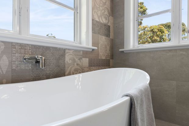 Close-up of a bright contemporary bathroom featuring a freestanding white bathtub beneath large white-framed windows. The walls are finished in patterned and stone-look grey tiles, with a wall-mounted chrome tap and a grey towel draped over the tub.