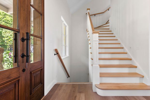 Bright residential entry hall featuring a dark timber glazed front door, white walls, warm timber flooring, and a white staircase with timber treads and handrails ascending to an upper level. The space has traditional detailing and abundant natural light.