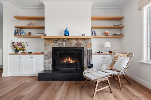 A bright, traditional living room centred on a stone-clad fireplace with a black firebox and raised hearth. White built-in cabinetry and open timber shelves flank the fireplace, displaying books and small decor. Two woven lounge chairs with cushions sit on warm timber flooring beside a window, creating a refined, inviting reading area.