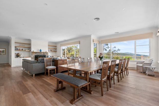 Bright open-plan living and dining room with large timber dining table, matching wooden chairs and bench seating, grey sofa, built-in shelving, fireplace, and expansive windows overlooking a green landscape. The room has warm timber flooring, white walls and ceiling, and a traditional yet relaxed residential interior.