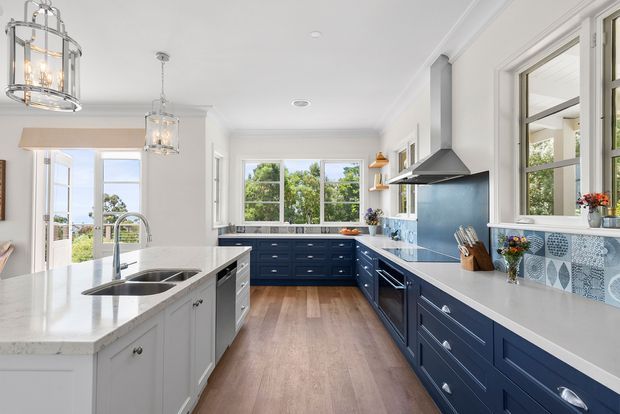 Bright, spacious kitchen with a large central island, navy-blue perimeter cabinetry, white island cabinetry and benchtops, patterned blue-and-white tiled splashback, timber-look flooring, pendant lights, and abundant natural light through multiple windows and glazed doors.