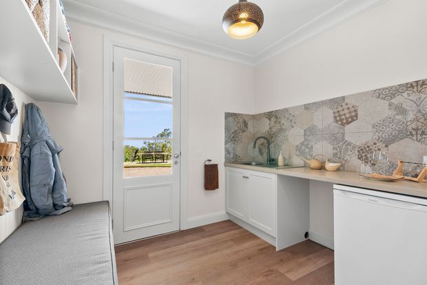 Bright, compact utility/laundry room with white cabinetry, a long pale benchtop, patterned geometric tile splashback, integrated sink, timber-look flooring, wall shelving, and a built-in bench with coat storage. A glazed exterior door provides natural light and views outside. A dark pendant light and simple cornices add understated character.