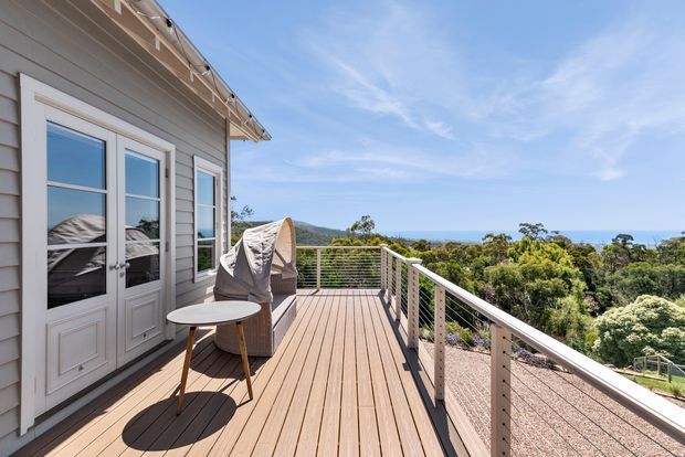A bright elevated outdoor timber deck or balcony attached to a weatherboard-clad home, with French-style glazed doors, a covered outdoor chair, small round table, and slim metal cable railing. The deck overlooks dense coastal vegetation and a distant blue ocean beneath a clear sky.