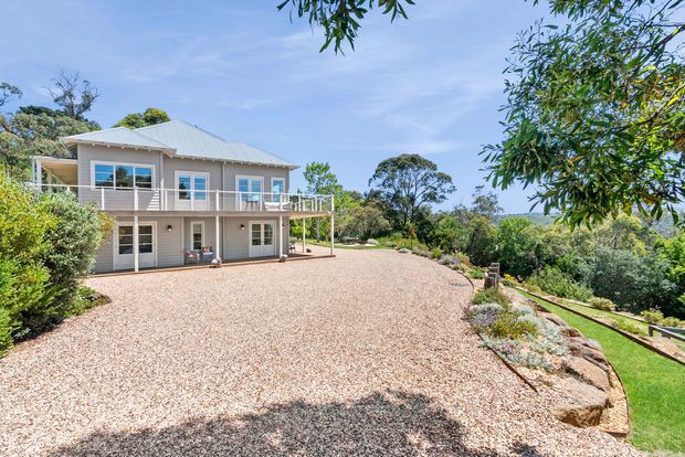A light-coloured two-storey residential house set within a landscaped garden. The home has a pale grey metal hipped roof, cream weatherboard-style exterior walls, white-framed windows and doors, and a wraparound upper-level balcony or veranda with simple metal railings. A broad gravel driveway or forecourt curves around the house, with planted garden beds, natural rocks and mature trees surrounding the property.