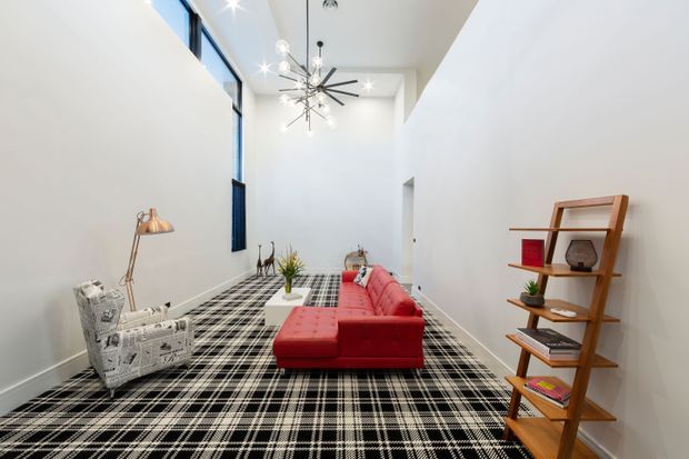 Bright, tall living room with white walls and a high ceiling, featuring floor-to-ceiling windows, a striking black starburst pendant light, a red sectional sofa, patterned armchair, glass coffee table, timber open shelving, and a bold black-and-white plaid carpet. The room has a spacious, gallery-like contemporary character with strong graphic contrasts.