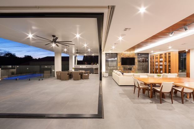 A spacious contemporary open-plan living, dining and outdoor entertaining area with fully retractable glazed doors. The interior features a large white sectional sofa, timber dining table and chairs, extensive warm timber built-in shelving, a stone-clad fireplace wall, and polished grey flooring. The covered outdoor terrace continues the same flooring and includes lounge seating, a dining setting, an outdoor kitchen or bar, a ceiling fan, and views toward a swimming pool. Recessed lighting and warm timber ceiling detailing create a refined indoor-outdoor setting.