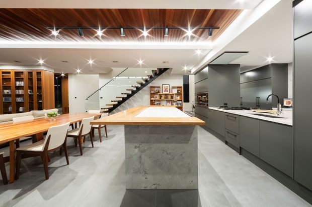 A spacious open-plan contemporary kitchen and dining area with a large central island, dark grey cabinetry, pale stone surfaces, and integrated appliances. A timber dining table and built-in timber shelving sit beside the kitchen, while a glass-sided staircase leads to the upper level. The room features a dramatic timber-lined recessed ceiling with exposed linear detailing and multiple recessed spotlights, creating a refined, high-end interior.