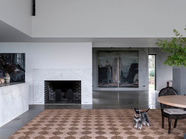 A refined contemporary living room with a double-sided or oversized fireplace clad in pale marble, polished concrete flooring, and a large patterned area rug. The space has white walls, a high ceiling, broad open-plan proportions, dark metal-framed glazing or fireplace doors, and an adjacent dining table. A small dog is present in the room.