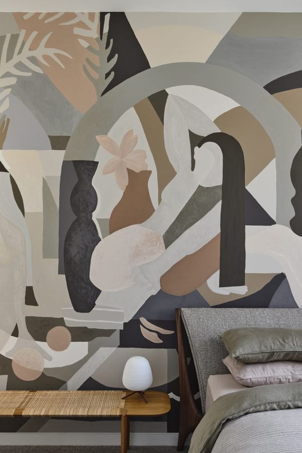 A styled bedroom corner featuring a large abstract mural with organic geometric forms in muted taupe, grey, cream, charcoal and blush tones. A textured upholstered bed with layered grey bedding occupies the foreground, accompanied by a low mid-century timber bench with woven cane seating and a small sculptural table lamp. The composition is calm, art-led and tactile.