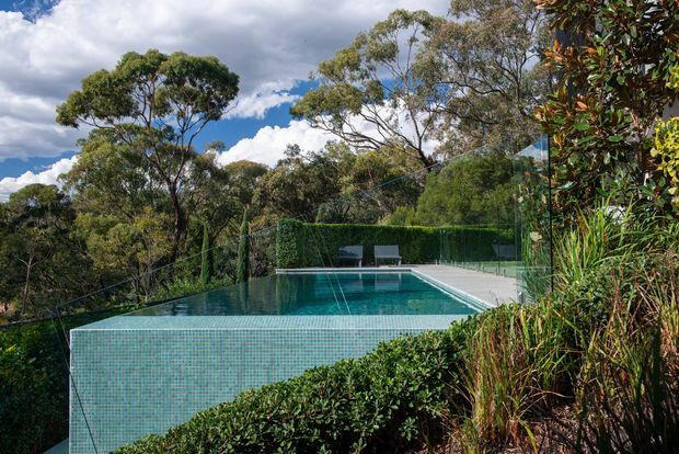 A landscaped outdoor swimming pool set into a wooded garden, with a turquoise mosaic-tiled infinity edge, glass safety fencing, pale paving, clipped hedges, and dense native-style planting. The scene is photographed outdoors beneath a partly cloudy sky.