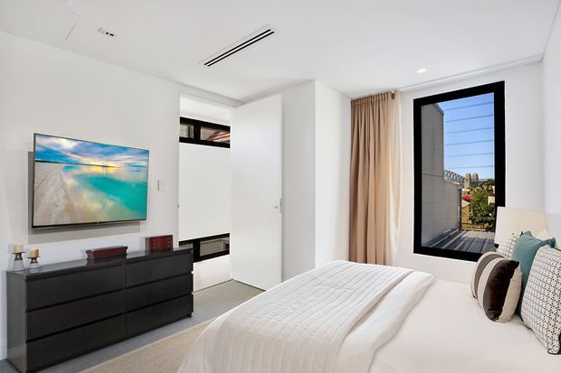 Bright contemporary bedroom with a large upholstered bed dressed in white bedding, a black six-drawer dresser with wall-mounted television, neutral carpet, full-height beige curtains, and a black-framed window. A partially open door reveals a compact adjoining area, while the room is finished with recessed lighting and a linear ceiling air vent.
