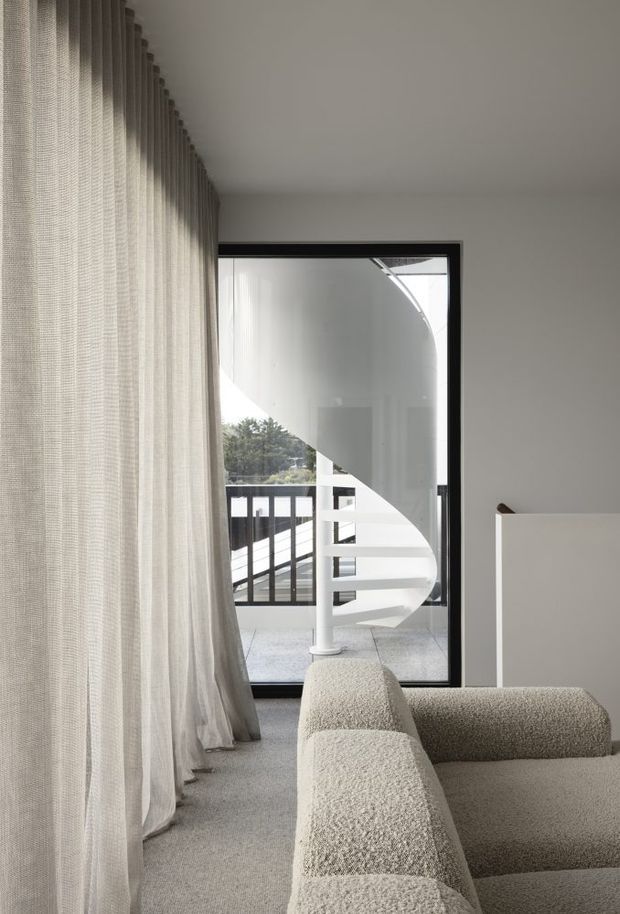 A softly lit, minimalist living room viewed toward a balcony through full-height glazing. Sheer textured curtains line the left wall, while a large cream boucle sectional sofa occupies the foreground. A white sculptural spiral staircase is visible beyond the black-framed glass door, with pale neutral walls and carpet creating a calm, refined interior.