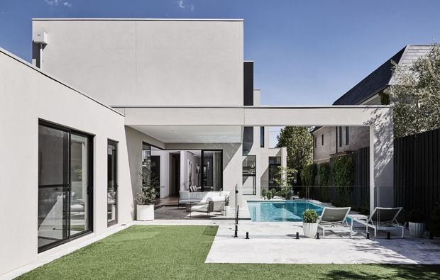 A modern courtyard-style residential exterior with crisp white rendered volumes, a covered outdoor passage, floor-to-ceiling glazing, a small swimming pool, pale paving, artificial lawn, and contemporary outdoor seating. Dark fencing and neighbouring pitched-roof homes frame the private outdoor space.